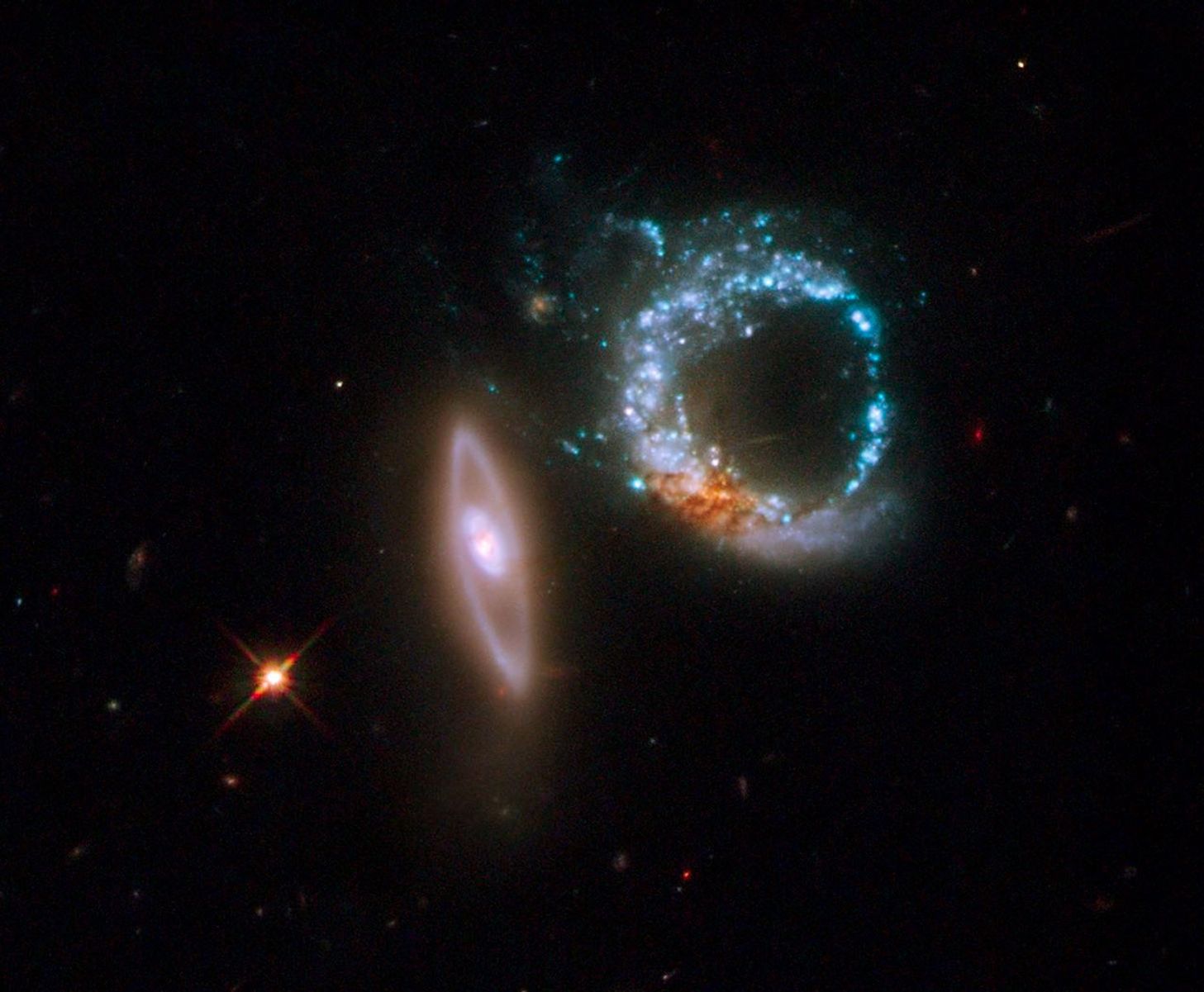 Left: a disk galaxy that appears nearly edge-on to our line of sight with its long axis vertical and just left of image center. It has a bright core with a smooth ring of starlight surrounding it. Right: a ring galaxy. Its pancake-shaped disk of material appears is a clumpy blue ring of intense star formation. A dusty reddish blob at the bottom of the blue ring may be the galaxy's original nucleus.